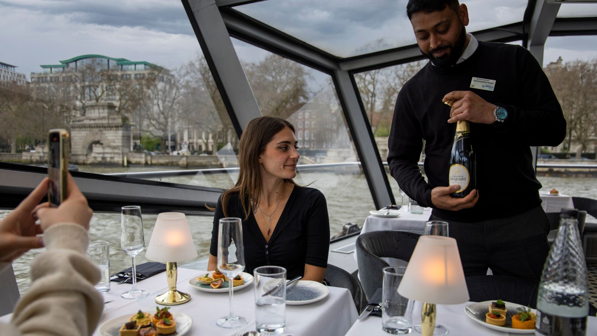 Enjoy entertainment and a live singer on board a London dinner cruise. Image courtesy of Golden Tours. Two people sitting at a table on a Thames cruise with a waiter holding a bottle of champagne.