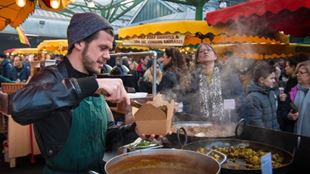 Ein Mann in einer grünen Schürze der Street Food von einer riesigen Pfanne in eine Essensbox für Kunden im Borough Market serviert.