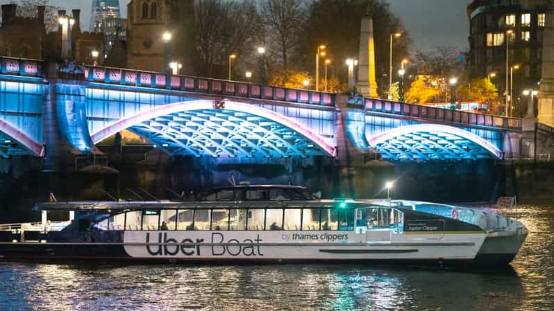 A black and white uber cruise boat sails underneath a London bridge lit up with blue lights
