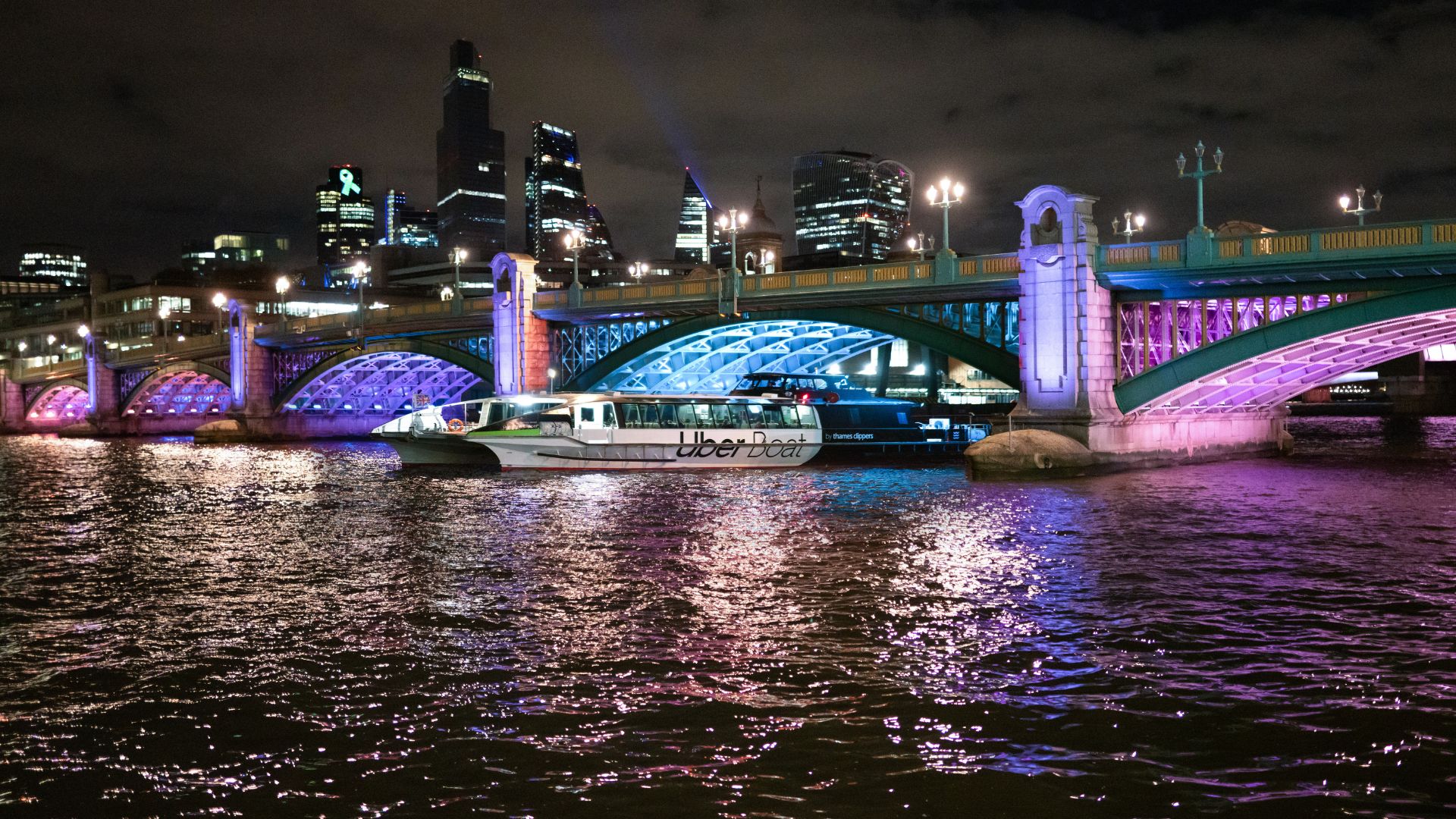 See London's bridges lit up at night on a river cruise. Image courtesy of Golden Tours. An Uber boat passes underneath a London bridge illuminated with pink, purple and blue lights.