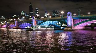 An Uber boat passes underneath a London bridge illuminated with pink, purple and blue lights.