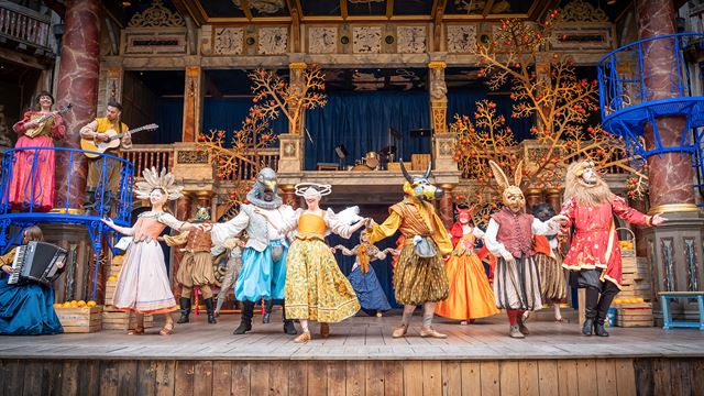 Actors wearing bright, colourful costumes and animal heads are dancing on the stage of the Shakespeare Globe Theatre.