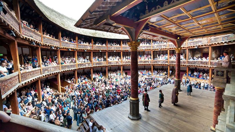 Experience a classic show at Shakespeare's Globe. Credit: Shakespeare's Globe. Image courtesy of the Shakespeare's Globe. Actors are standing on the large wooden stage at the Shakespeare Globe Theatre, facing a full auditorium.