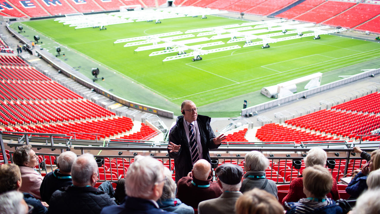 Découvrez les secrets du stade de Wembley en compagnie d'un guide expert. Image fournie par Golden Tours. Un guide parle en face d'un groupe assis dans les gradins du stade de Wembley, avec vue sur le terrain en contrebas.