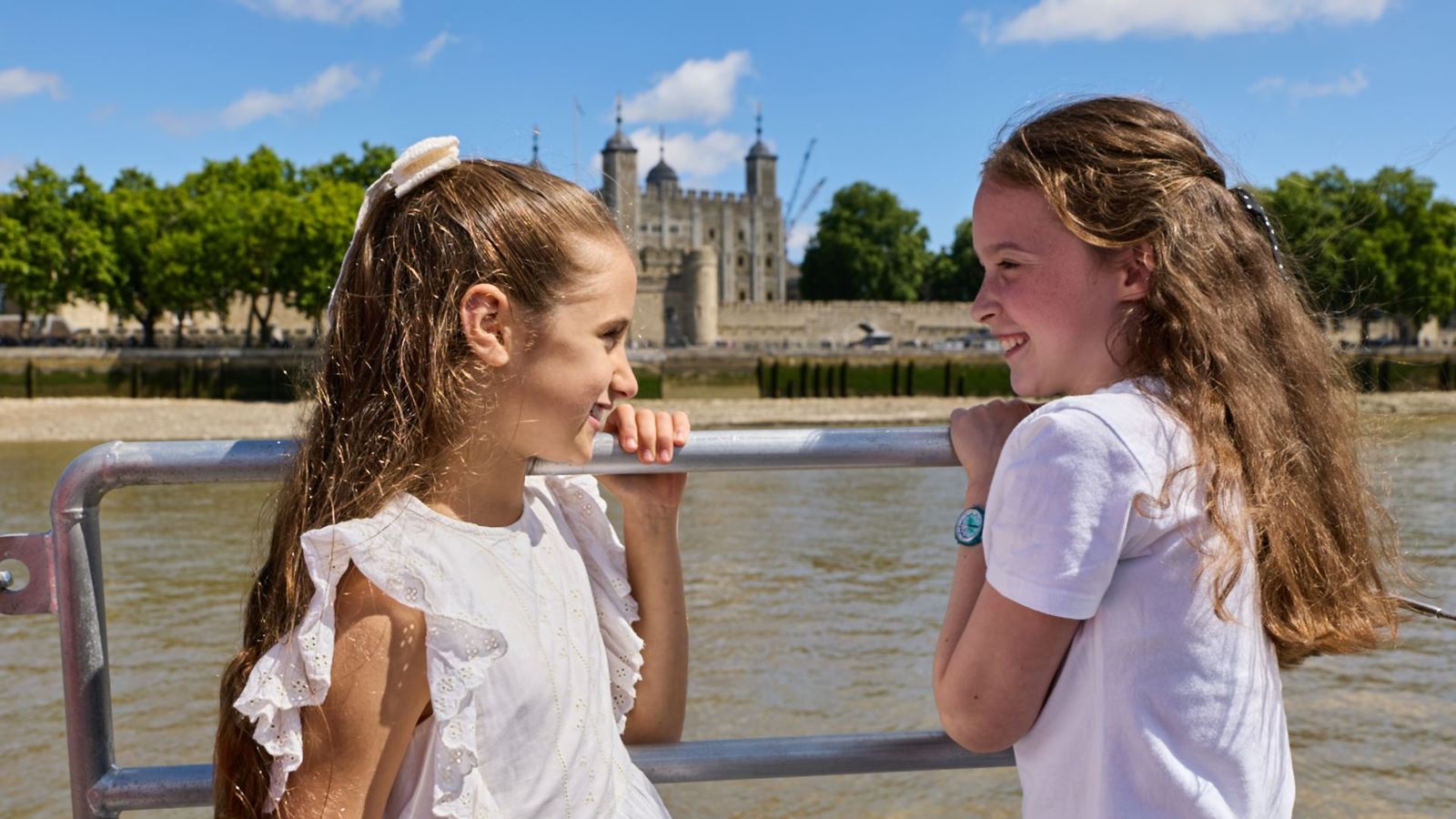 Two girls look towards each other aboard a river boat, with the Tower of London visible across the River Thames.