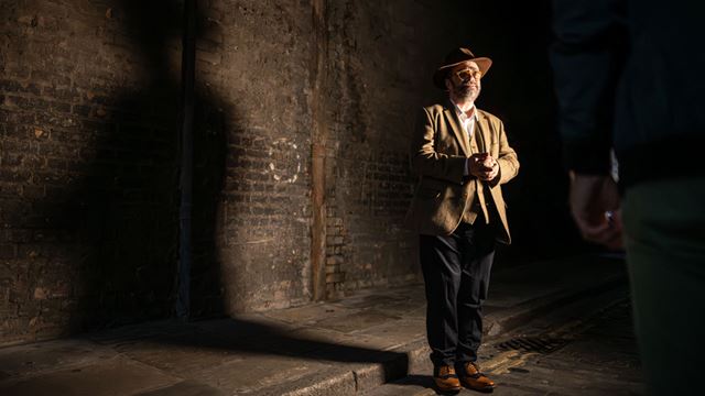 A tour guide dressed of a tweet suit and a brown hat is standing in a dark alleyway of East London during a Jack The Ripper walking tour. 