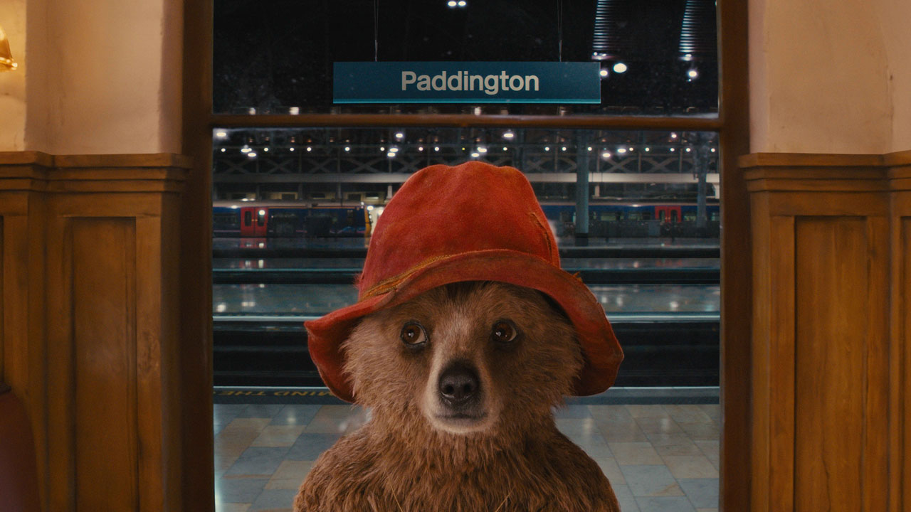 Suivez l'ours Paddington dans ses aventures à travers Londres lors d'une visite guidée. Image fournie par Golden Tours. L'ours Paddington porte un bob rouge et se tient a l'entrée de la gare de Paddington.