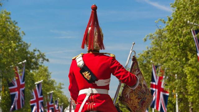 A  Royal Guard dressed in red sits on a horse and rides down The Mall, with Union Jack flags lining the road.