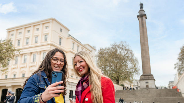 Zwei Frauen machen auf The Mall ein Selfie, die Straße die zum Buckingham Palace verläuft, mit dem Duke of York Monument im Hintergrund.