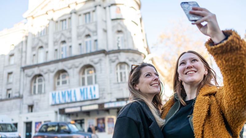 Join a Theatreland Walking Tour and hear about famous shows of the past and present. Image credit: London & Partners/ Michael Barrow. Two women taking a selfie in front of Mamma Mia theatre sign.