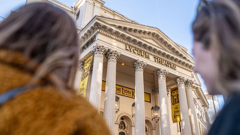 Two women stand in front of London's Lyceum Theatre, admiring the iconic venue that hosts Disney's top musical The Lion King.