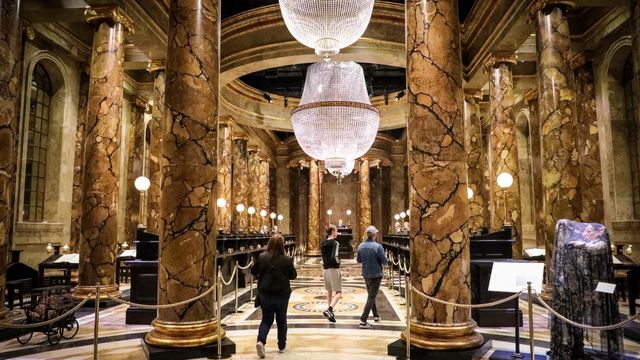Les gens se promènent dans la salle dorée de la banque Gringotts des studios Harry Potter, avec ses grands piliers et ses lustres étincelants.