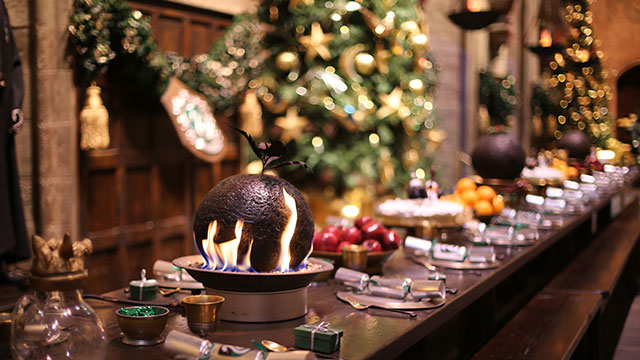 Weihnachtspudding ziert die Tische in der Großen Halle © Warner Bros. Studio Tour London – The Making of Harry Potter. Ein in Flammen stehender Weihnachtspudding auf einem langen, mit Tellern gedeckten Tisch vor einem mit goldenen Sternen geschmückten Weihnachtsbaum