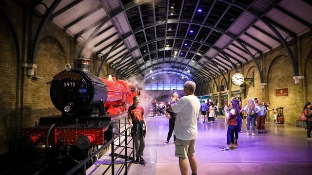 A tall man in grey takes a photo of a woman in a red jumper next to the red and black hogwarts express train at the warner bros studio tour.