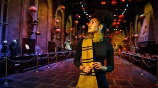 A person stands in the Great Hall of Hogwarts, decorated with floating jack-o'-lanterns for Halloween. The hall features long tables set for a feast, stone walls with gothic architecture, and statues of winged creatures. The person is wearing a yellow and black striped scarf and looking to the side, capturing the magical ambiance