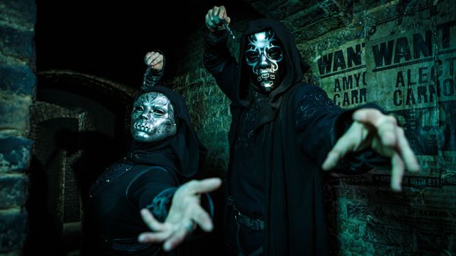 Two people in dark hooded robes and metallic masks with intricate designs pose dramatically in a dimly lit, stone-walled setting. Their hands are extended forward as if casting spells. Behind them are old, tattered 'WANTED' posters with partially visible names, creating a mysterious and eerie atmosphere.