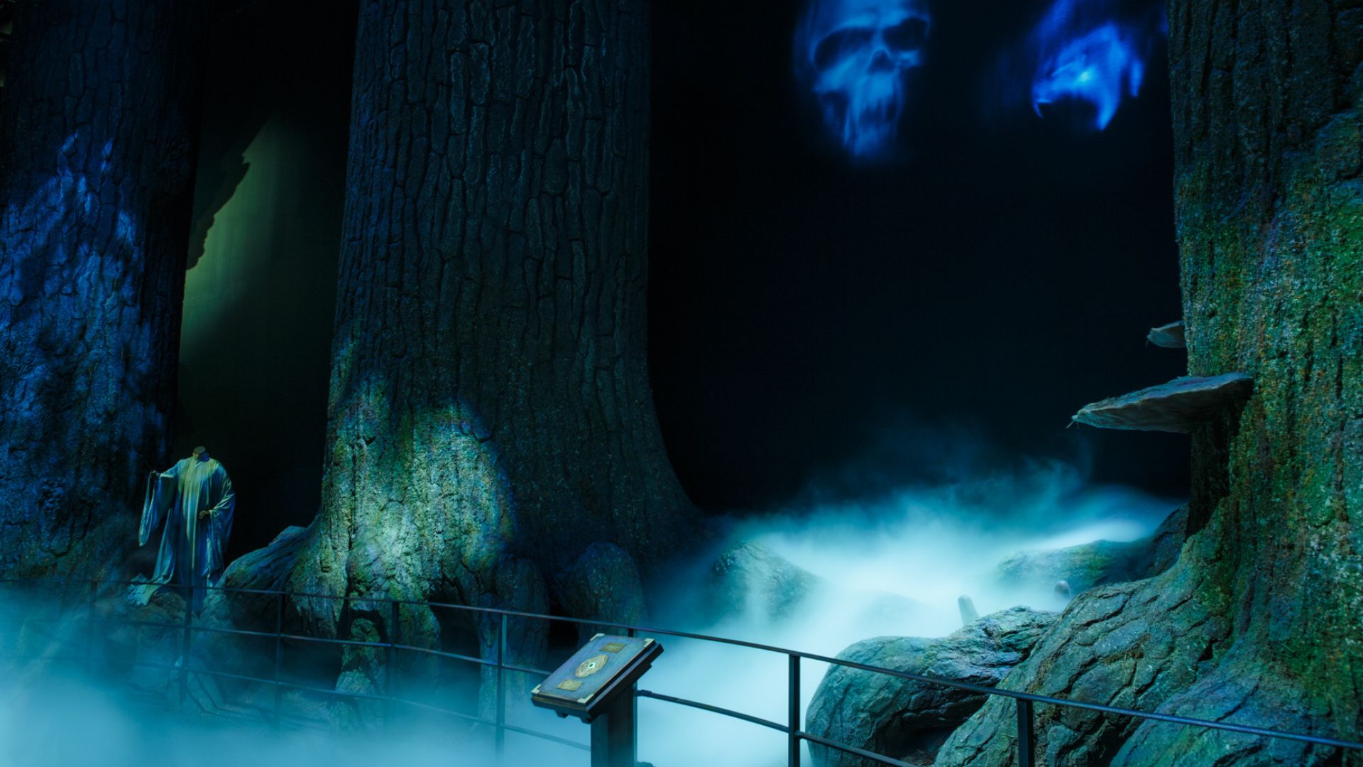 Venture into the Forbidden Forest at Warner Bros. Studio. Image courtesy of Golden Tours. A dark, eerie forest scene with large tree trunks and mist on the ground. A headless statue in a flowing robe stands to the left, illuminated by blue light. Ghostly blue skulls float on the right. In the foreground, there is a metal railing and an information plaque, creating a mysterious and haunting atmosphere.