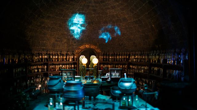 A dimly lit, arched room resembling a potion laboratory, with shelves of glass bottles and jars. Cauldrons and scientific instruments sit on tables in the center, illuminated by two glowing orbs. Ghostly blue images of a skull and a hand are projected onto the stone wall above, creating an eerie atmosphere.