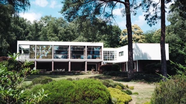 View of concrete and glass modernist house, The Homewood, and surrounding garden and trees.