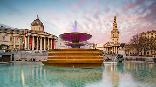 A fountain lit up in Trafalgar Square with the National Gallery behind.