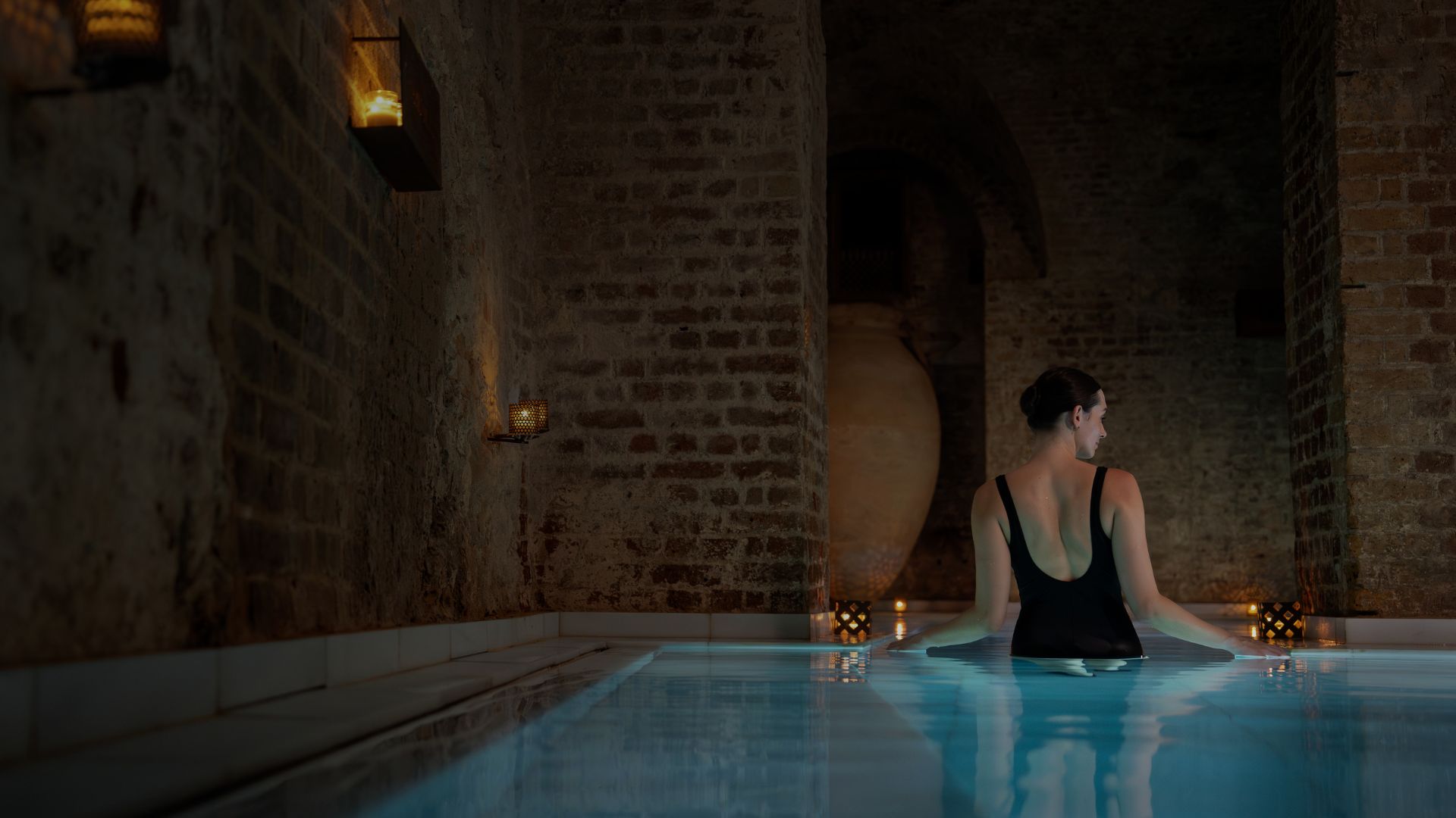 Person relaxing in a candlelit indoor pool with stone walls at a spa bathhouse.