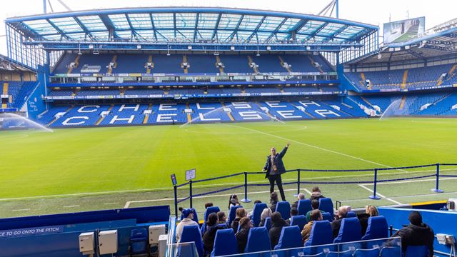Guide pointing to the stadium pitchside at Chelsea Football Club with a group of people sitting on seats in front of them.