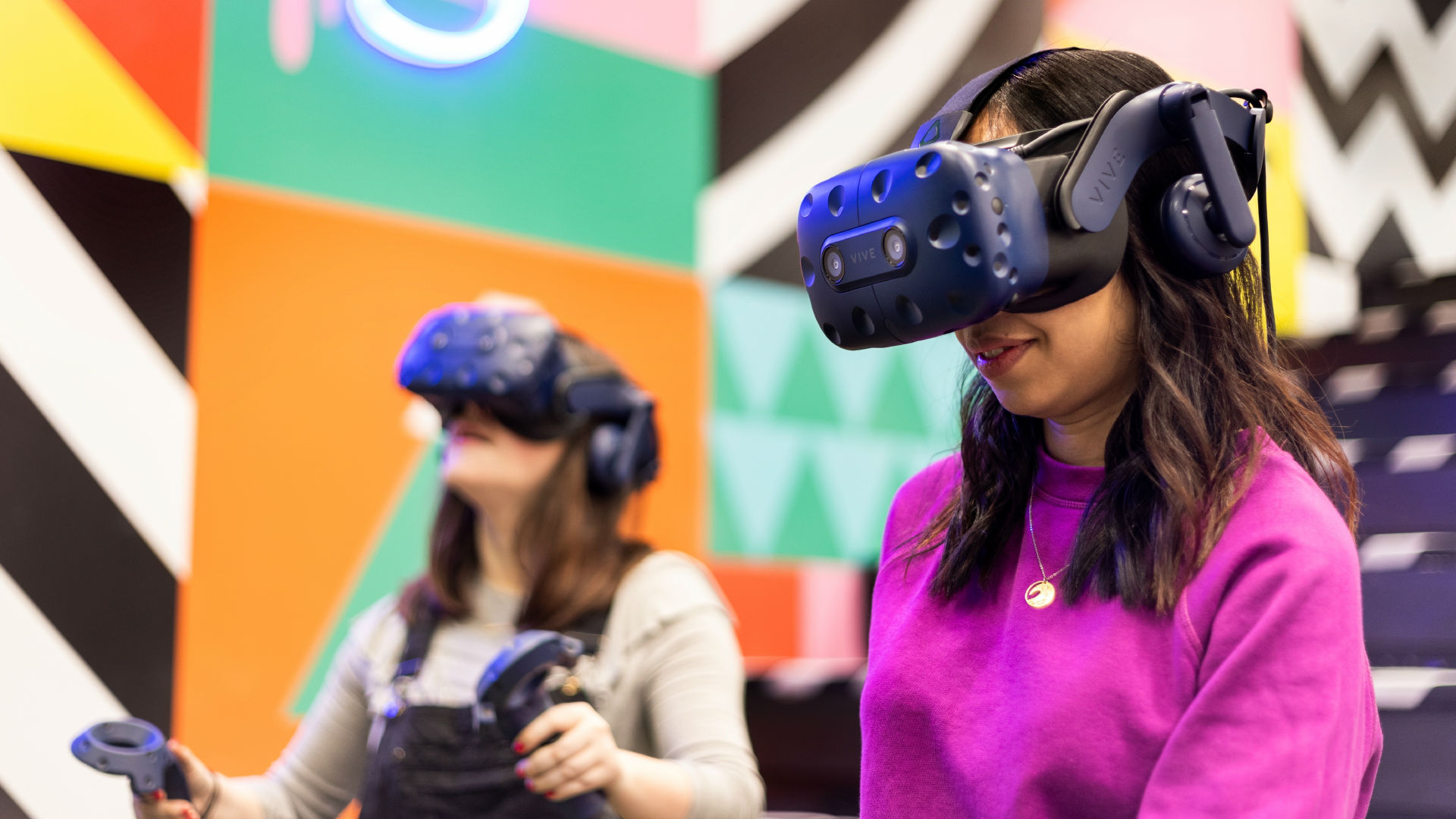 Two girls test out VR headsets in a colourfully decorated room. 