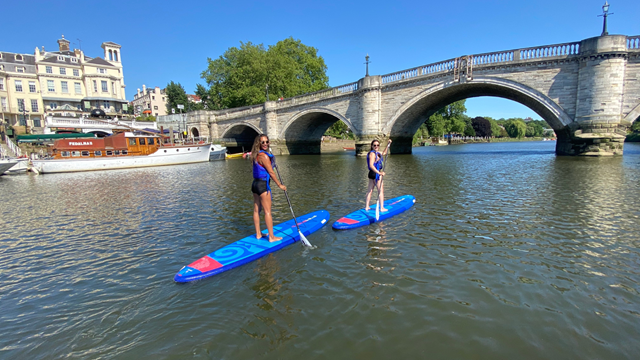 Paddleboarder on the river Thames beneath a railway bridge in Richmond, with a red train crossing above.