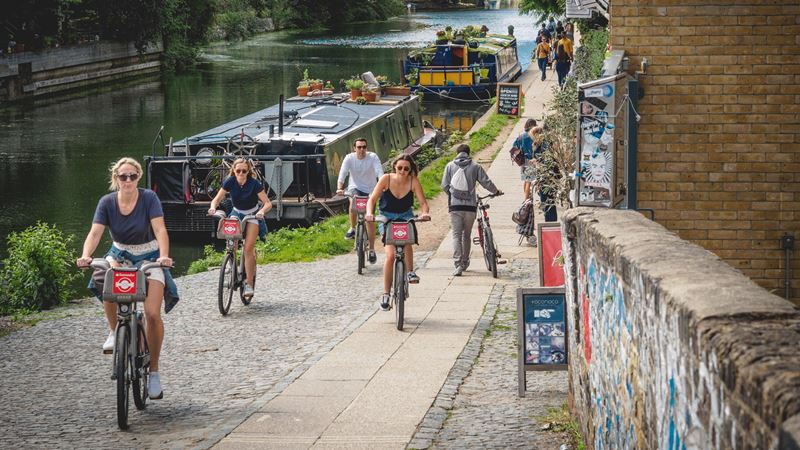 Explore the Thames Path by bike. Image courtesy of Shutterstock. A group of friends cycle next to the City Basin canal on Santander bikes with boats on the river in the background