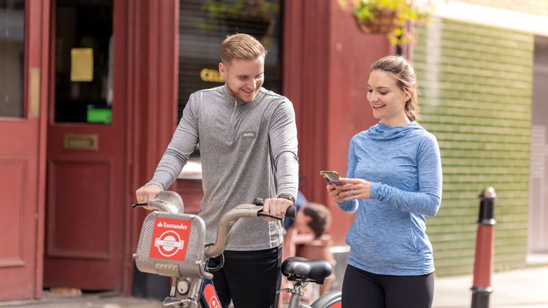 A man and woman walk along a street smiling at each other while the woman is on her phone and the man pushes a Santander bicycle