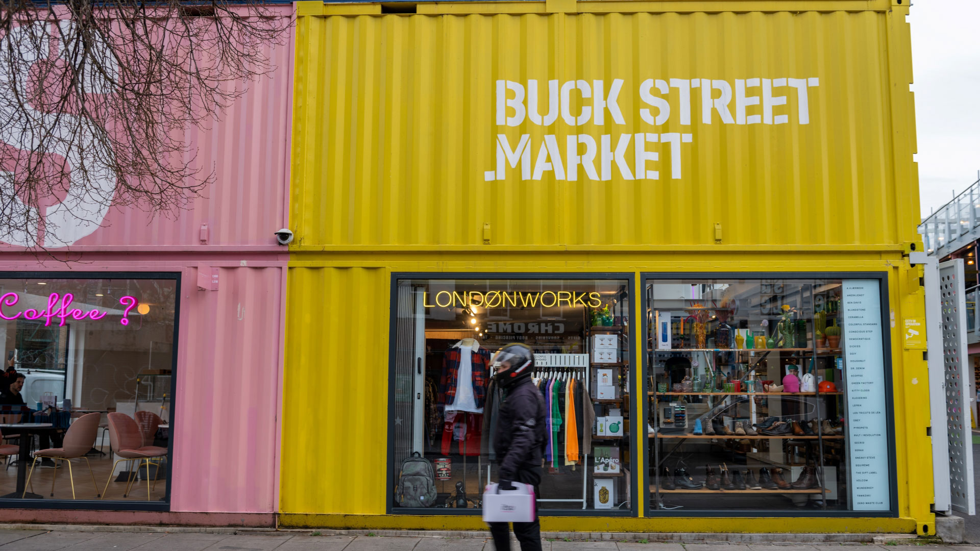 Man walking past a bright pink and yellow shipping container with shops inside which has been converted into Buck Street Market 