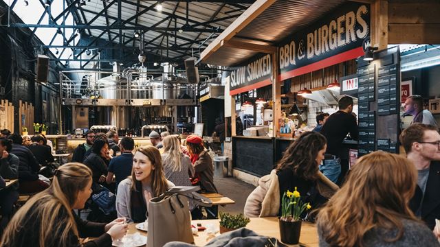 Benches with groups of people sat chatting and enjoying food at an indoor market with different food stalls cooking fresh food