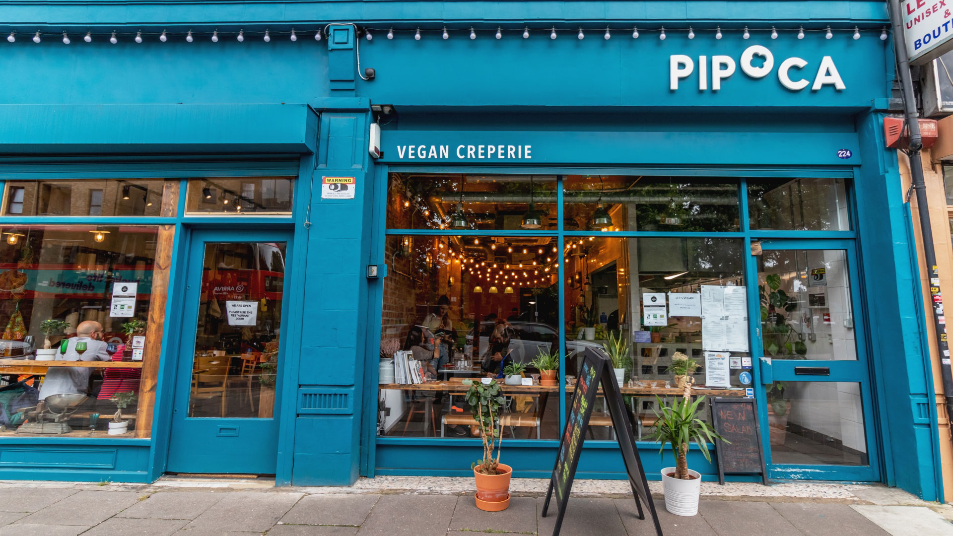 The blue painted Pipoca storefront in Brixton with people sat inside at the creperie surrounded by plants and fairy lights