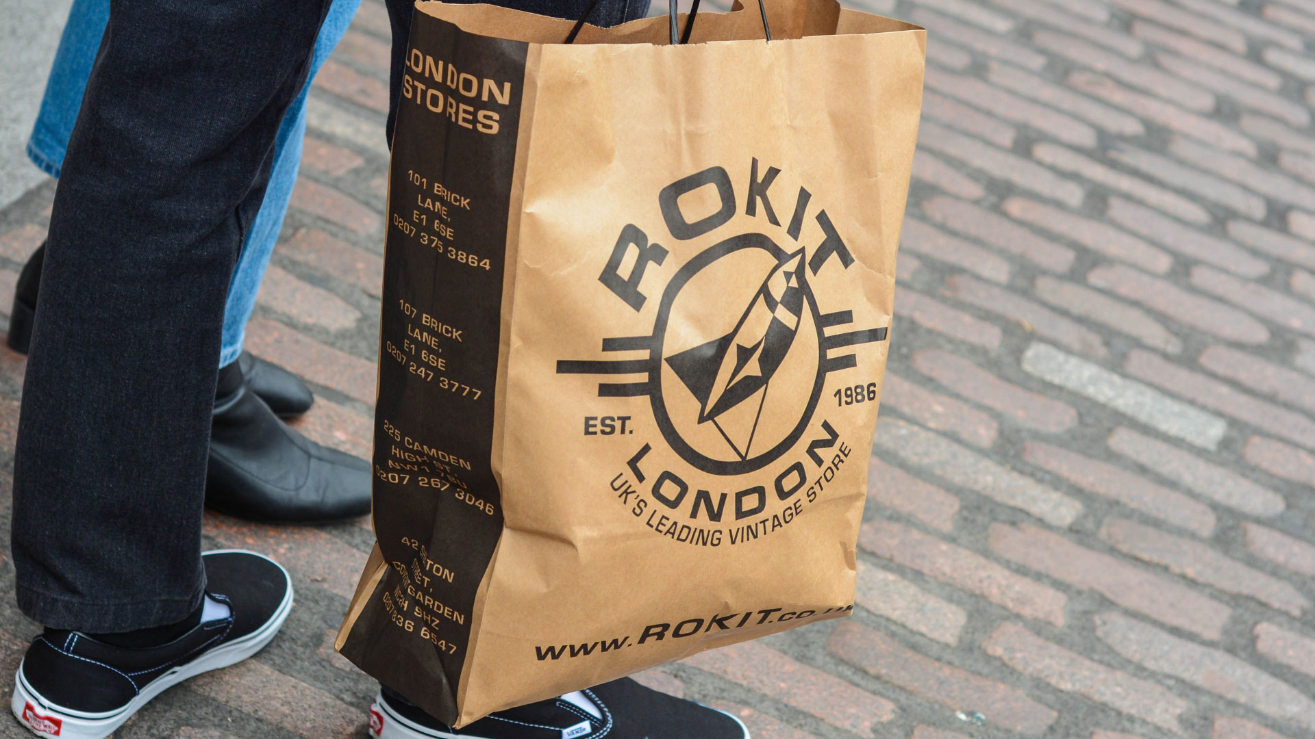 Someone holding a brown paper bag with the Rokit London logo on the front wearing jeans and black vans shoes