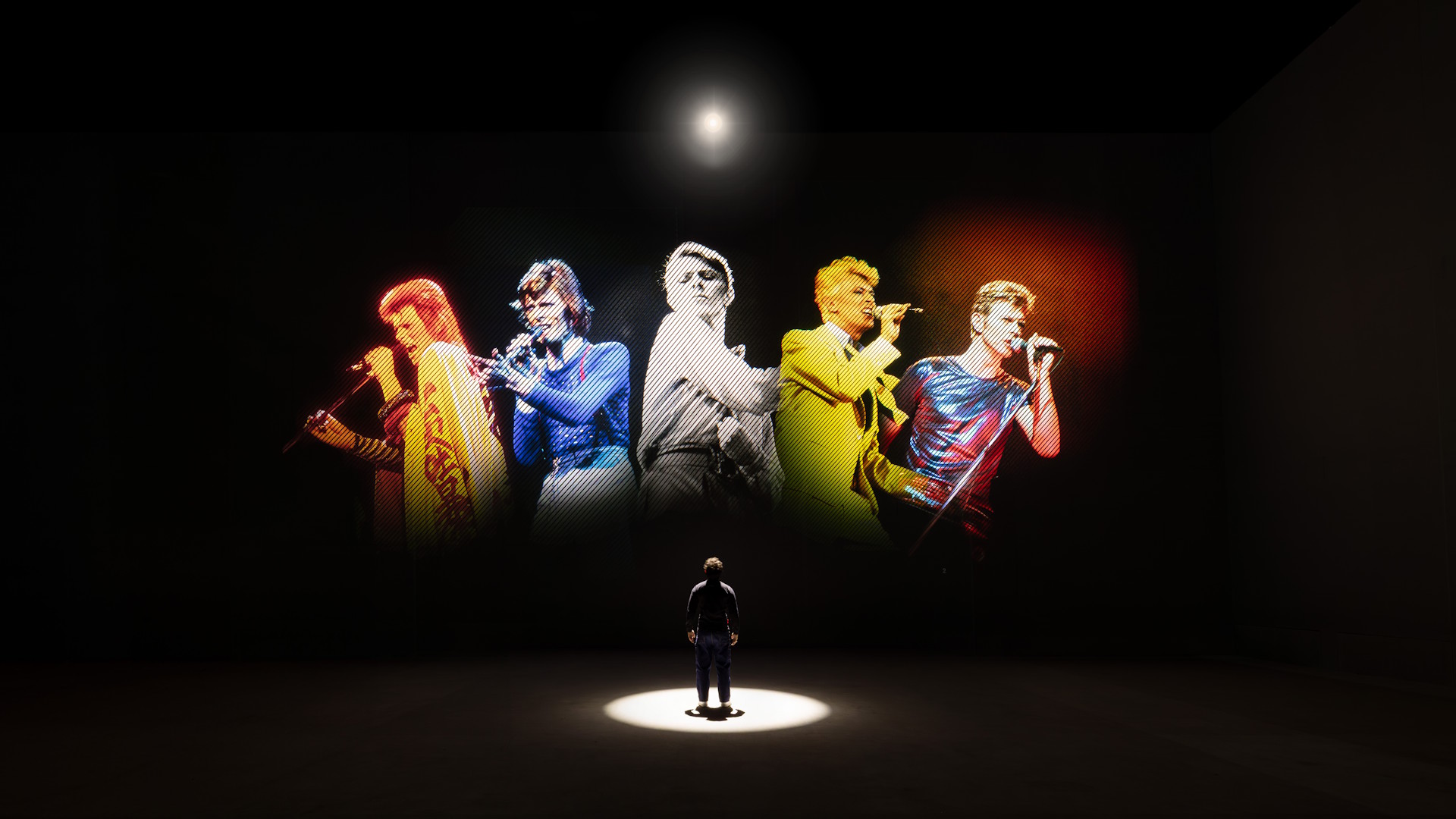 A person stands in a dark room with a light shining down, infront of a black screen with various images of David Bowie holding a microphone.