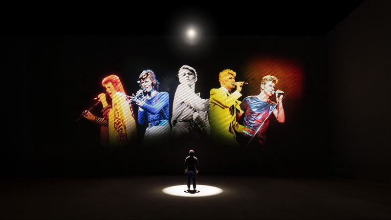 A person stands in a dark room with a light shining down, infront of a black screen with various images of David Bowie holding a microphone.