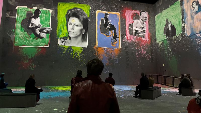 Go beyond the performer with audio clips and interviews as part of David Bowie: You're Not Alone at Lightroom. Image courtesy of Jonny Payne. Six images of David Bowie surrounded by various colours on the screens at Lightroom, with audience members sitting in front of the screens.