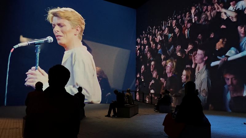 Feel as though you're part of a Bowie gig with footage from iconic concerts in this immersive show. Image courtesy of Jonny Payne. Lightroom's large screens showing David Bowie with a microphone on stage, and concert goers on a side screen, as part of the show David Bowie: You're Not Alone.