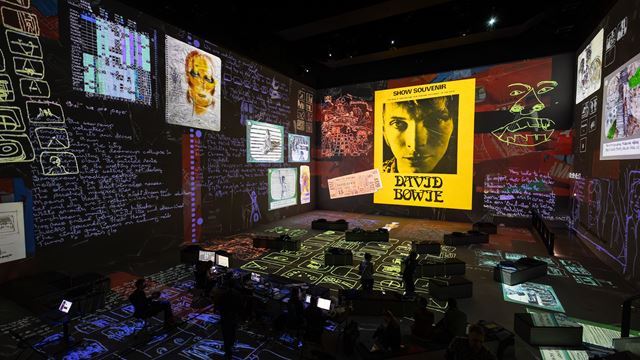 The darkened interior of Lightroom showing various archival material about David Bowie, and people sitting on the floor looking up at the floor-to-ceiling screens.