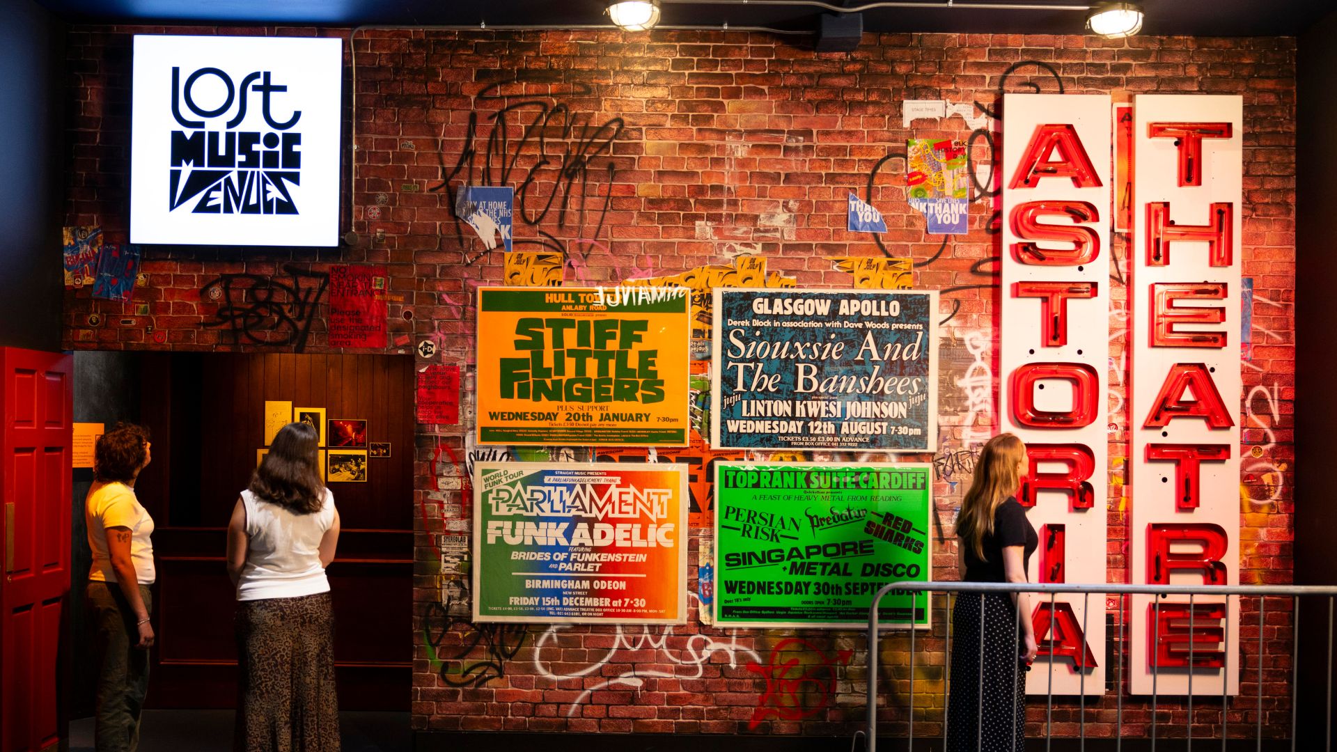 Explore the Lost Music Venues exhibition at the V&A, discovering iconic gig posters, venue signage and the stories behind some of Britain's most influential music spaces. Image courtesy of V&A. Photo credit: David Parry. Visitors explore the Lost Music Venues exhibition at the V&A, standing in front of a graffiti-covered brick wall displaying iconic gig posters and illuminated theatre signage. The immersive display recreates the atmosphere of Britain's historic live music venues, with bold colours, vintage artwork and memorabilia celebrating the country's music heritage.