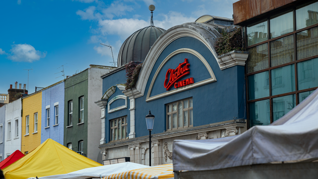 A photo of the light blue front exterior of Electric Cinema in Portobello with its trademark red neon logo and Portobello Road market stalls in the foreground