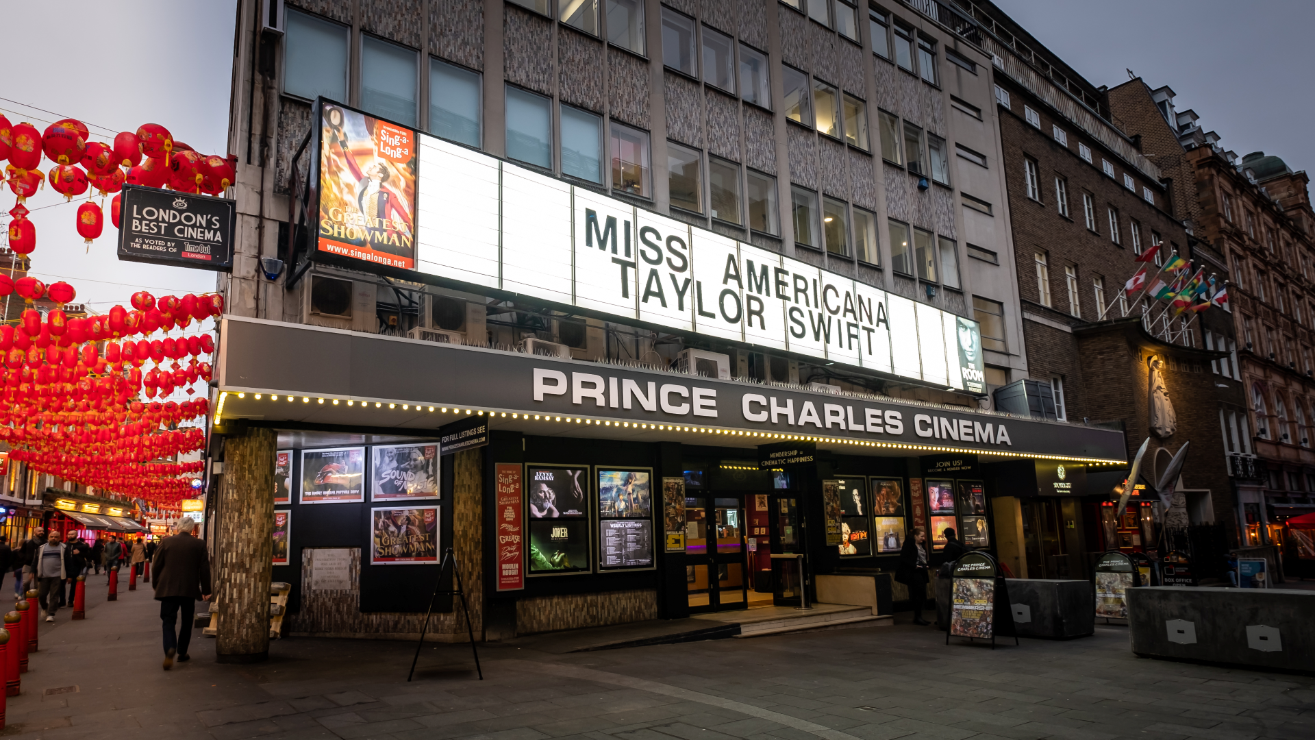 A photo of the front exterior of Prince Charles Cinema in Chinatown with a retro backlighted board with the films showing and red Chinese lanterns lining the street in the background