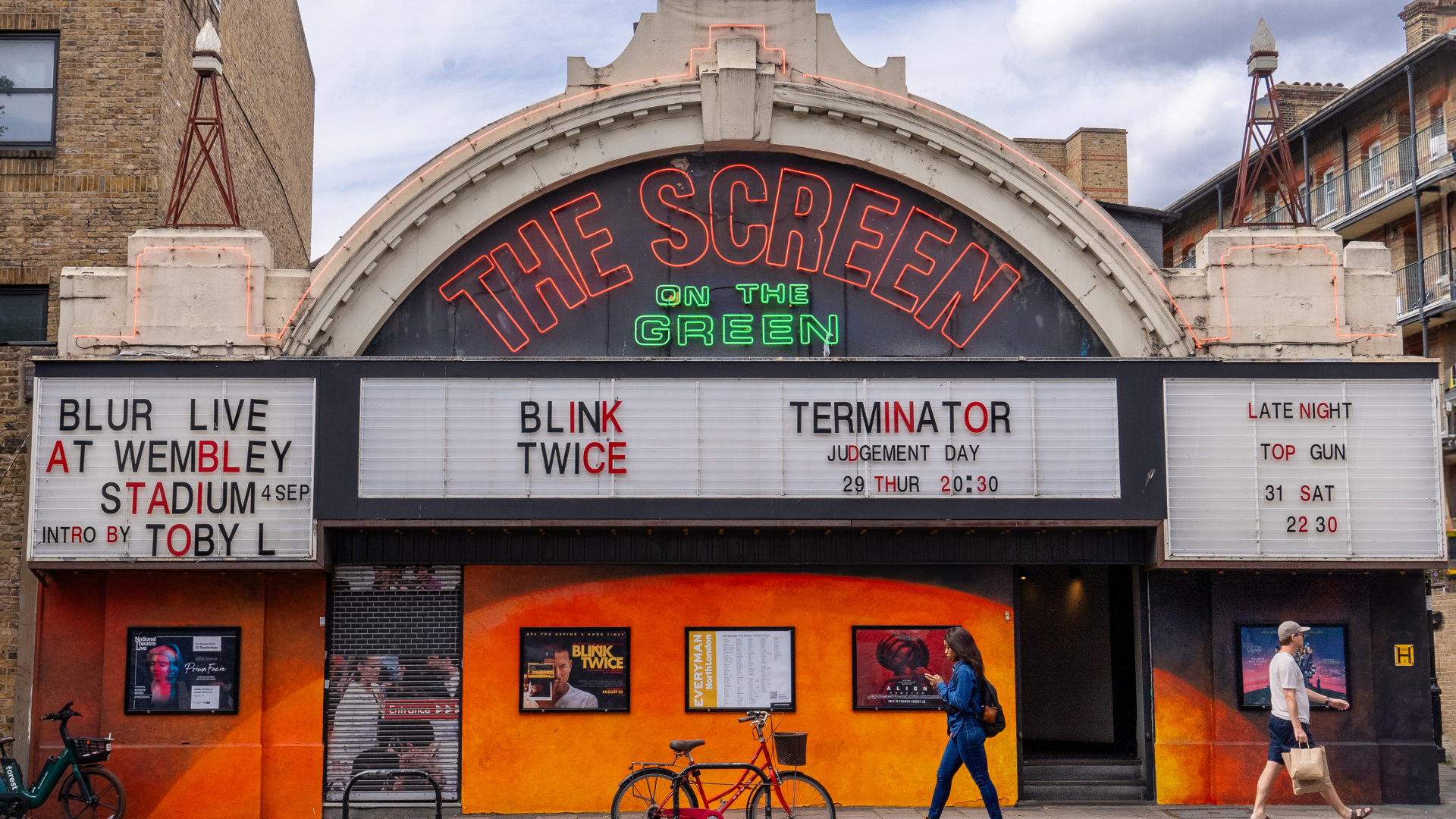 A photo of the front exterior of Screen on the Green with its red and green neon logo and retro white boards spelling out the names of films on show as people walk passed in the foreground