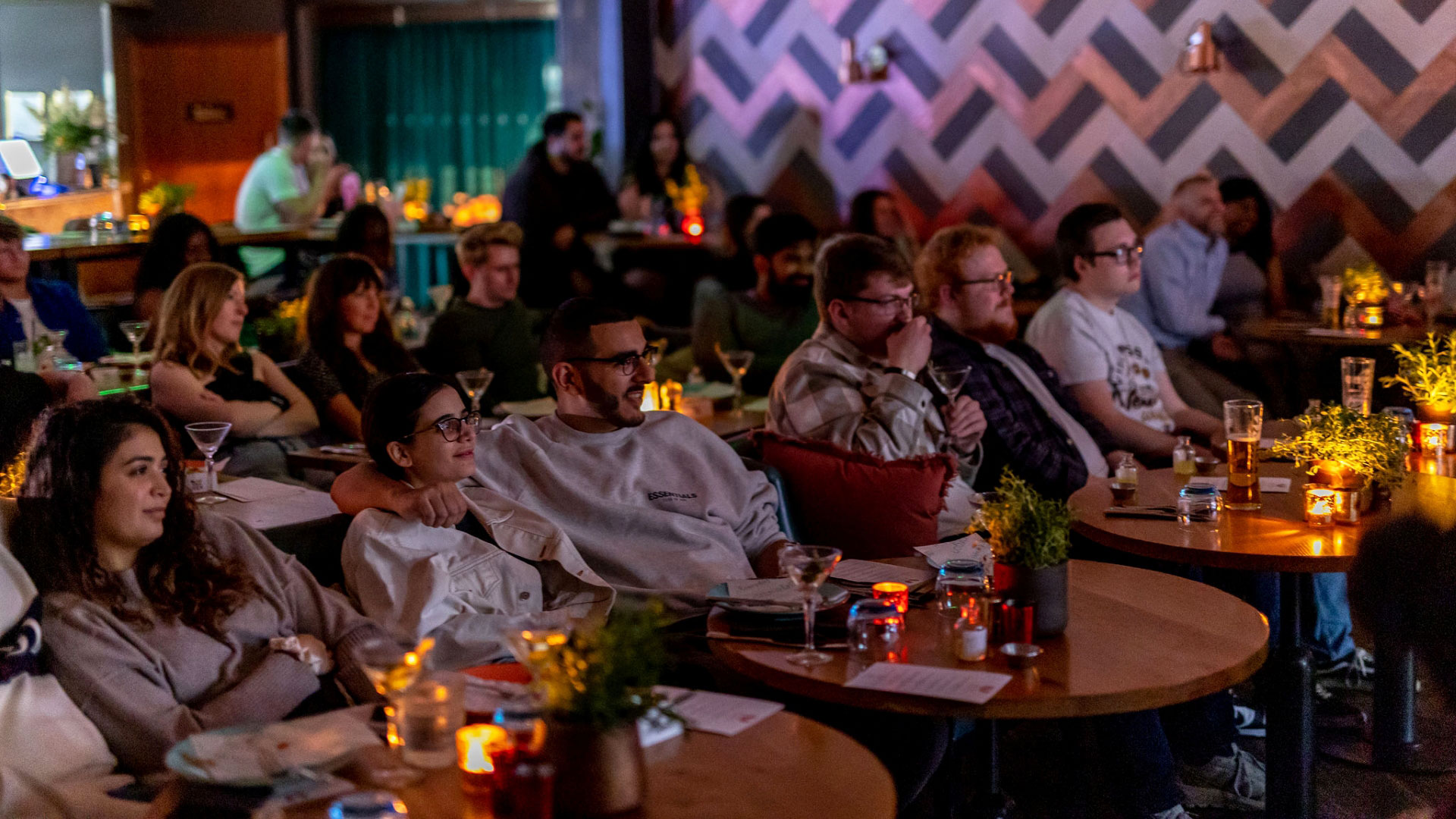An audience of people sit in comfy chairs looking forward at a film screen with tables in front of them with lamps and drinks ready to be served food 