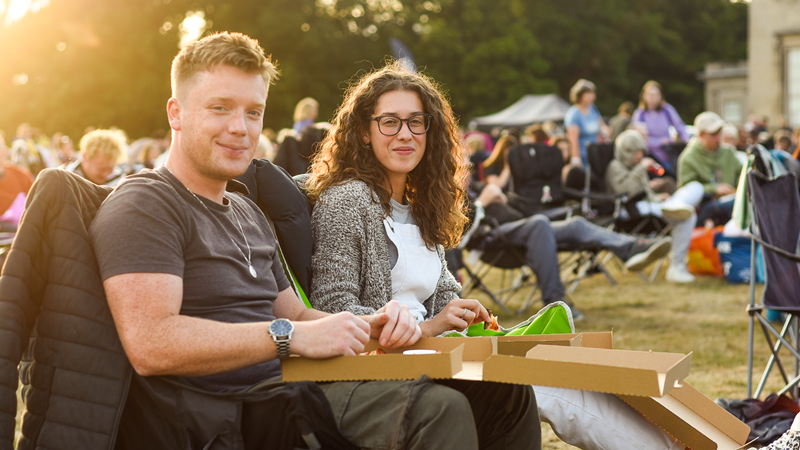 A couple smile at the camera whilst sitting on comfy chairs as the sun goes down behind them and they hold a pizza box on their laps surrounded by other revellers at an outdoor cinema screen