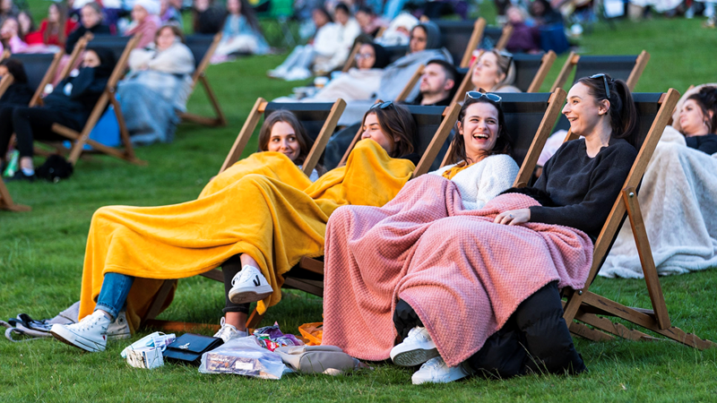 Grab some mates and get ready to watch a film in these unique outdoor cinemas in London. Credit: Adventure Cinema. Image courtesy of Adventure Cinema. Four girls sit on deck chairs on grass in a park surrounded by other revellers with blankets over them ready to watch a film at an outdoor cinema screen
