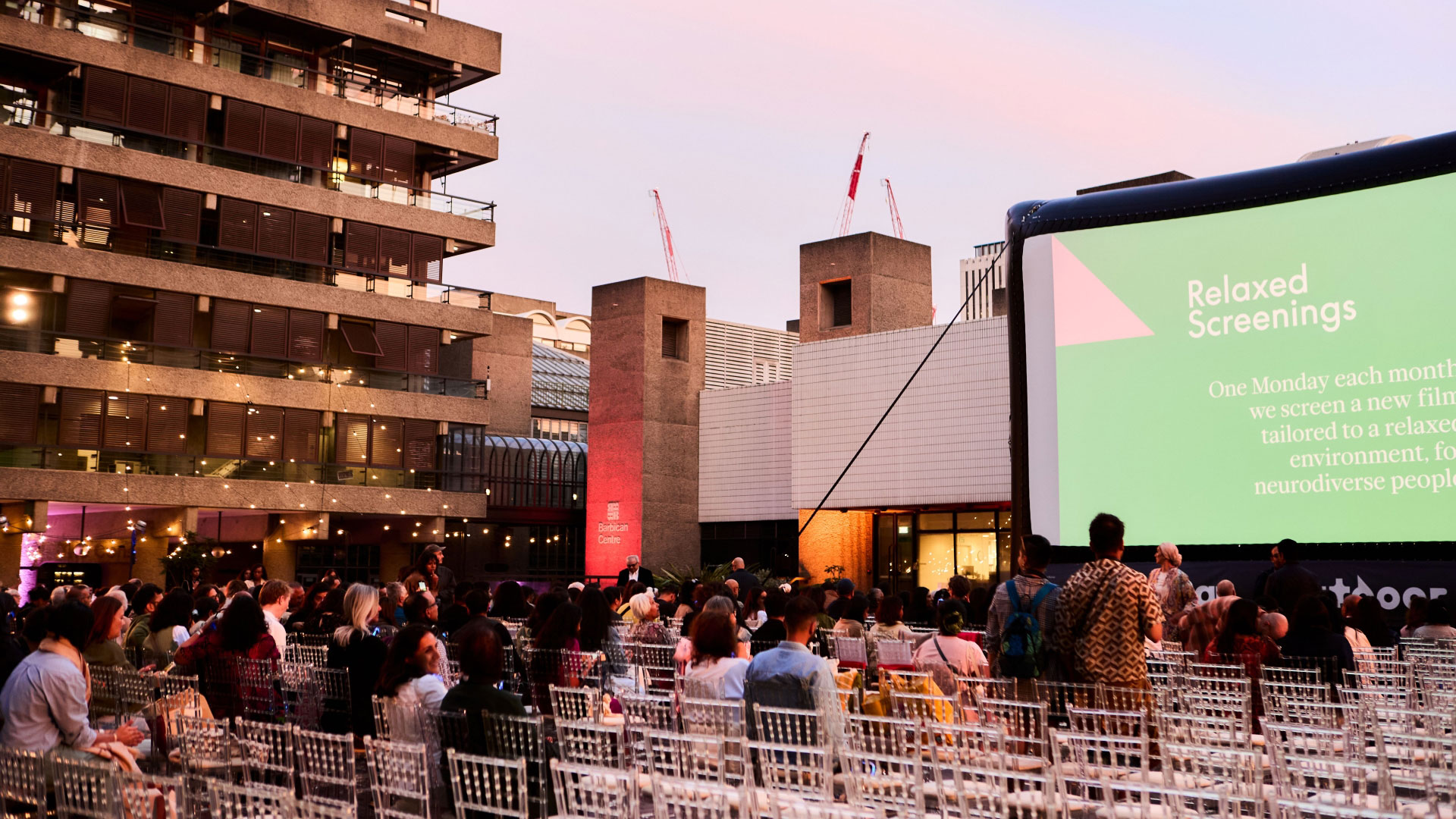 A photo of seats in front of a popup cinema screen in Barbican's Sculpture Court as people sit and walk to their seats with Brutalist buildings in the background