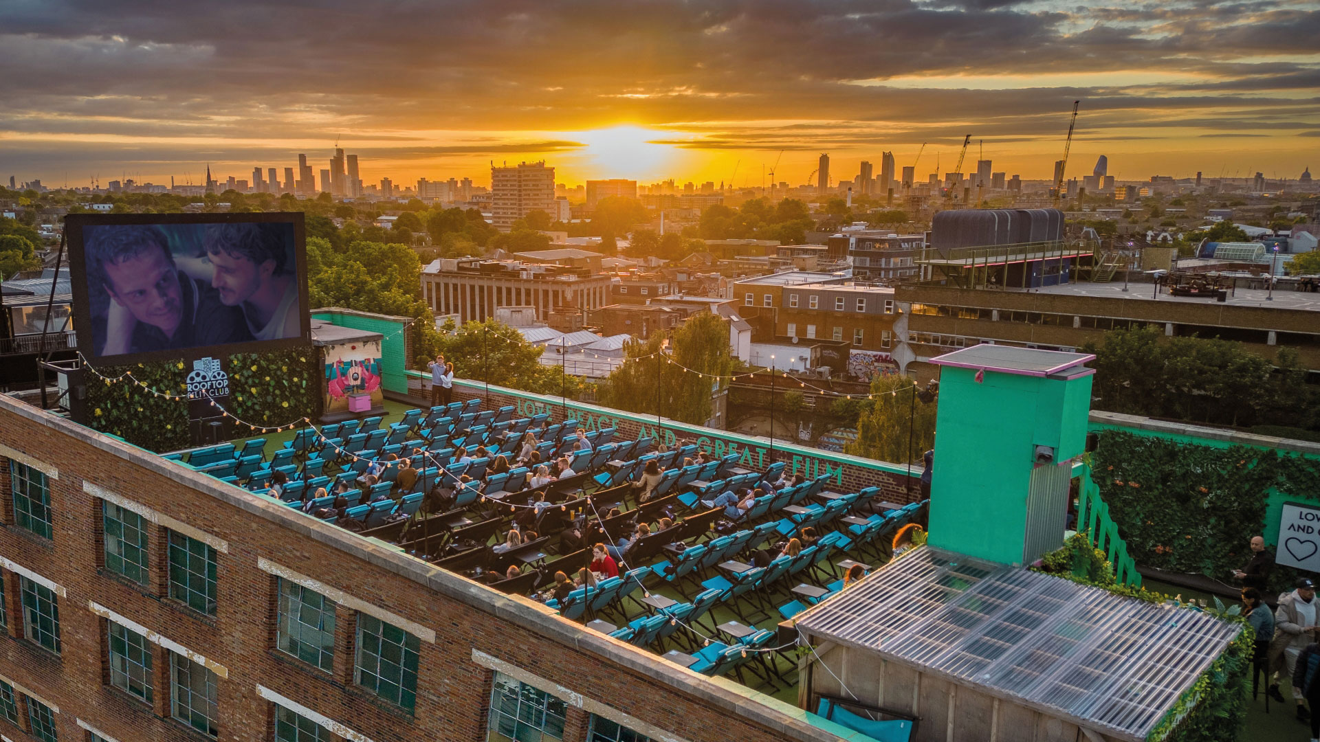 A photo from above Rooftop Film Club in Peckham with the screen playing a movie to seats of people on the rooftop as the sun sets behind London's buildings in the distance