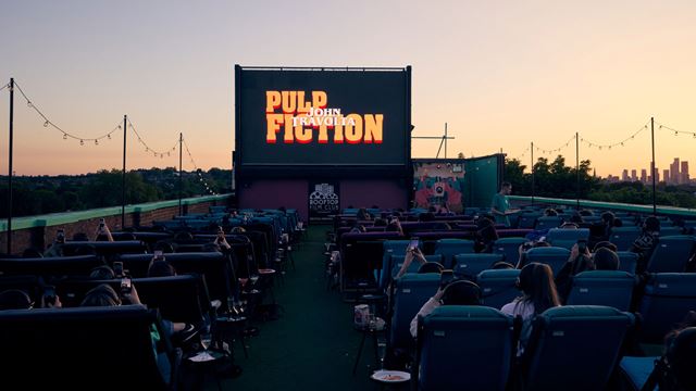 A photo of Rooftop Film Club Peckham from behind the seats of people with the screen showing "Pulp Fiction" title screen as the sun sets in the background