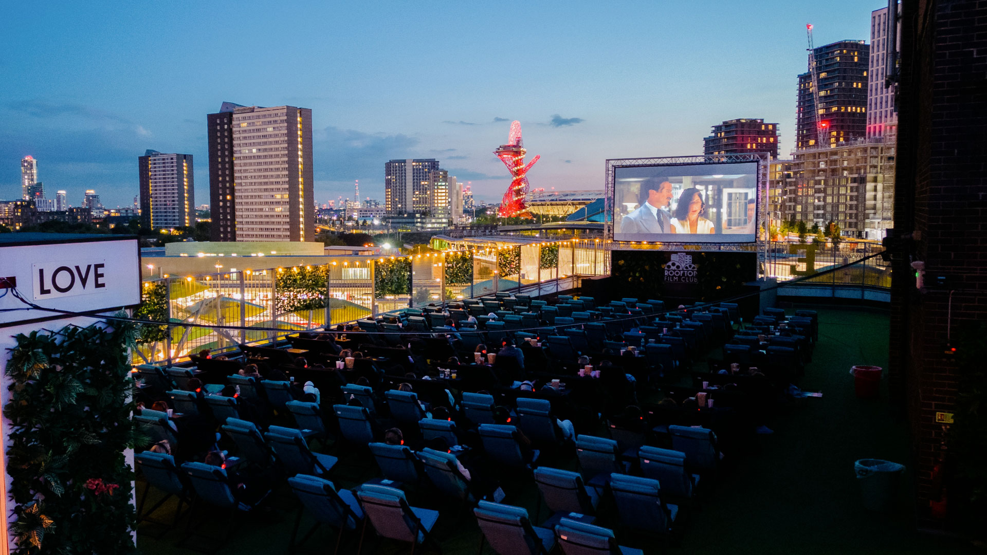 A photo from above Rooftop Film Club in Stratford with the screen playing a movie to seats of people as London's skyline creates a backdrop in the distance